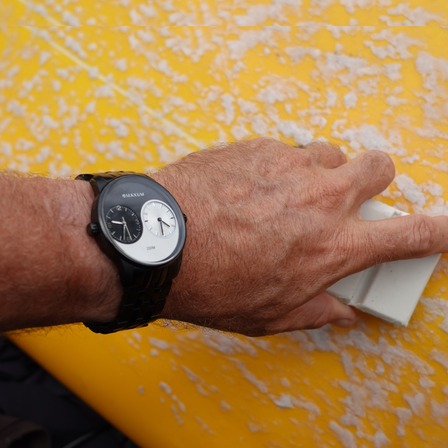 Maxum x Taylor Steele Collab analogue watch with a black stainless steel bracelet, black case, and a matte black dial featuring ying yang design and a dual timekeeping system. Hand wearing watch putting wax on a yellow textured surfboard