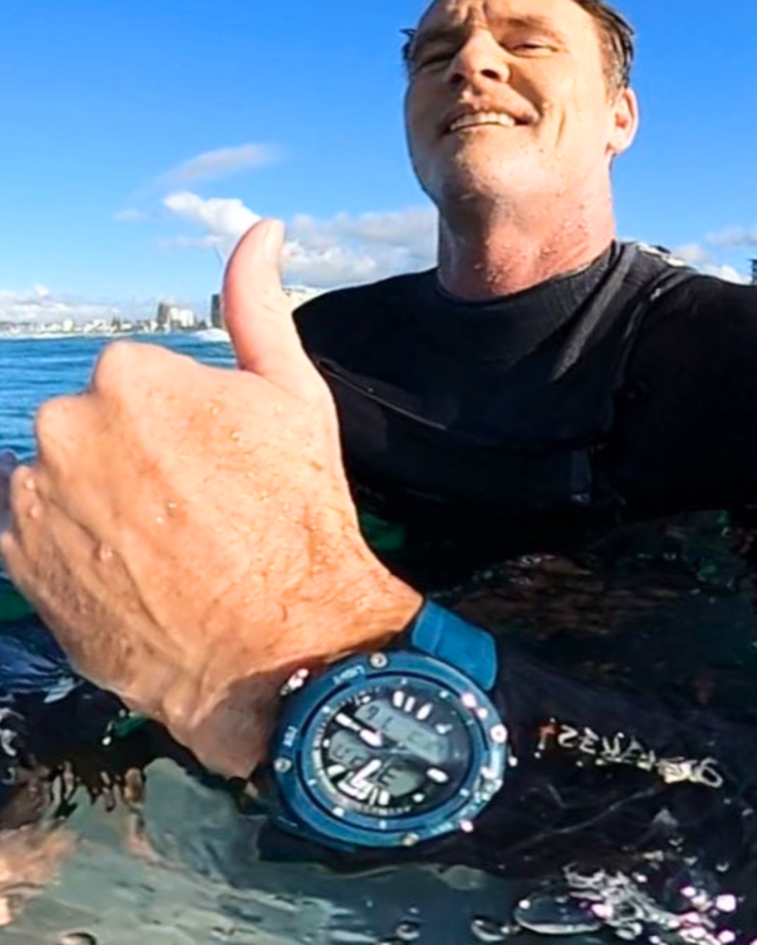 Maxum blue Conquest Analogue Digital Watch with a silicone strap and a digital display. Person in wetsuit with blue watch in water, city skyline in background