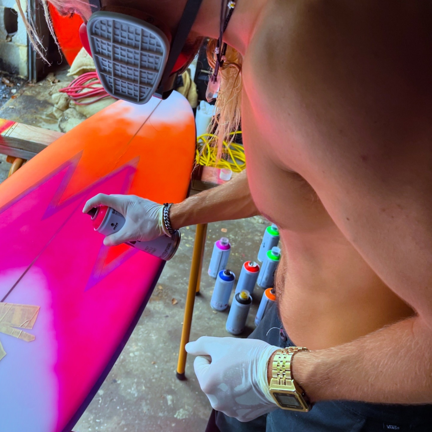 Kyuss King wearing Raglan Gold Tide Watch with a black dial and a stainless steel gold-tone bracelet. The watch has a square case with a digital display showing the time and tide features. Kyuss King is spray painting a colourful surfboard with various spray paint cans in the background.
