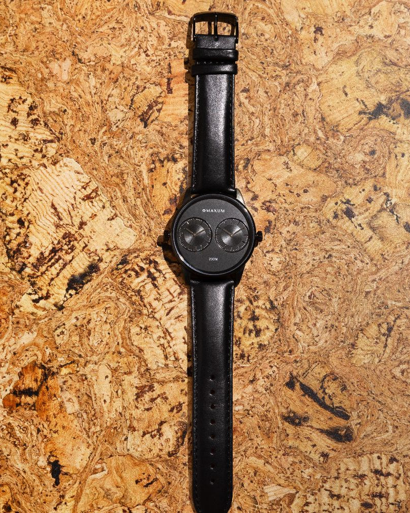 Maxum x Taylor Steele collab- black analog watch with a black dial, black case, and black leather band, branded with 'MAXUM' on the top and indicating '200M' water resistance. On cork backround