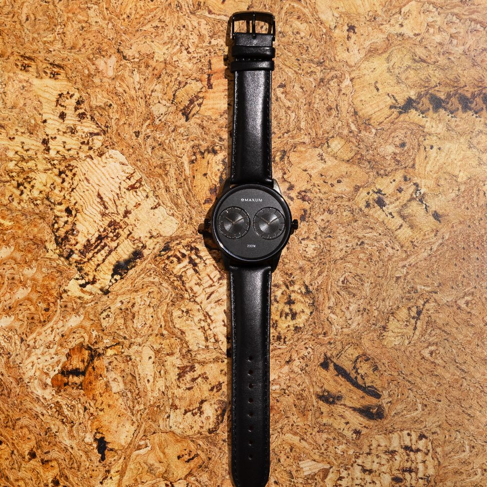 Maxum x Taylor Steele collab- black analog watch with a black dial, black case, and black leather band, branded with 'MAXUM' on the top and indicating '200M' water resistance. On cork backround