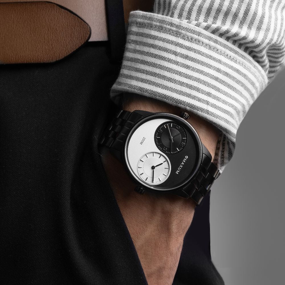Maxum x Taylor Steele Collab analogue watch with a black stainless steel bracelet, black case, and a matte black dial featuring ying yang design and a dual timekeeping system. On wrist with shirt striped shirt cuff and tailored black pants