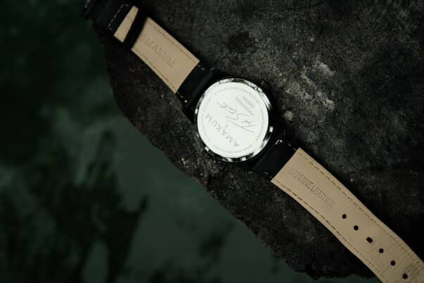 Maxum x Taylor Steele collab- black analog watch with a black dial, black case, and black leather band, back view on black rock surrounded by green water