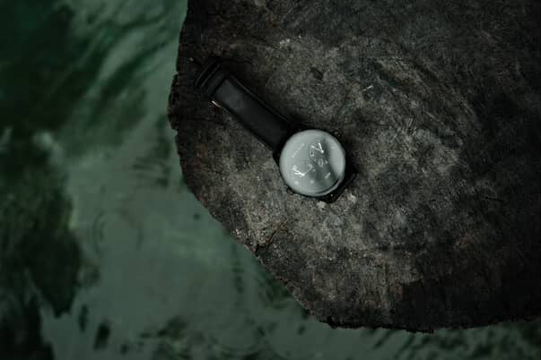 Maxum x Taylor Steele collab- black analog watch with a black dial, black case, and black leather band, on a black rock surrounded by green water