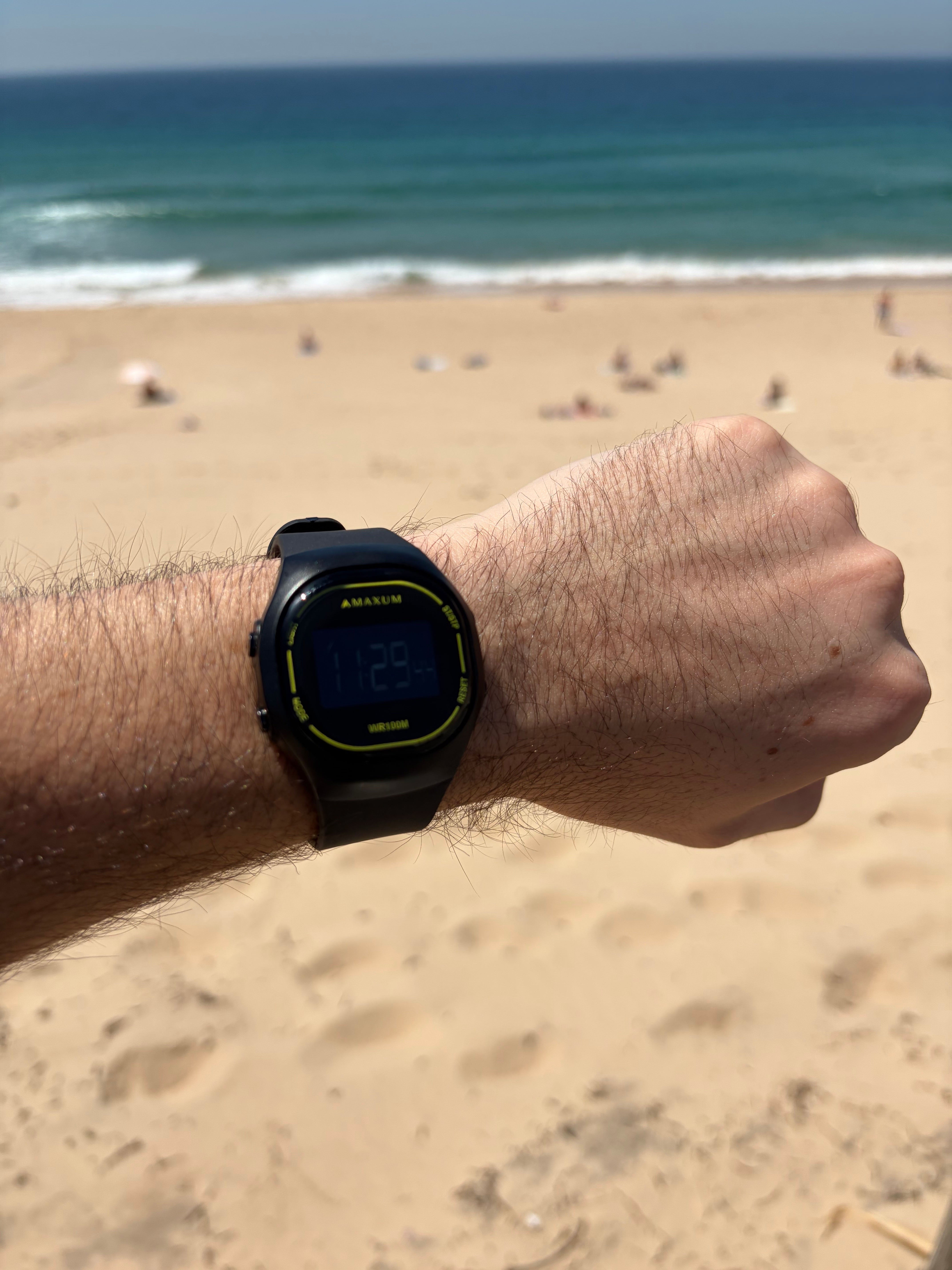 Maxum Nomad digital watch with a black case and black band, featuring yellow accents and a digital display showing the time. Person wearing a smartwatch on a beach with ocean in the background