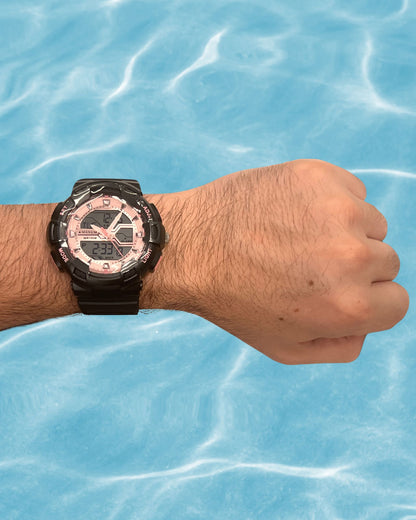 Hand wearing a A black and pink Maxum brand analog-digital watch with a resin strap and buckle closure, featuring a digital display and multiple watch features against a blue water background