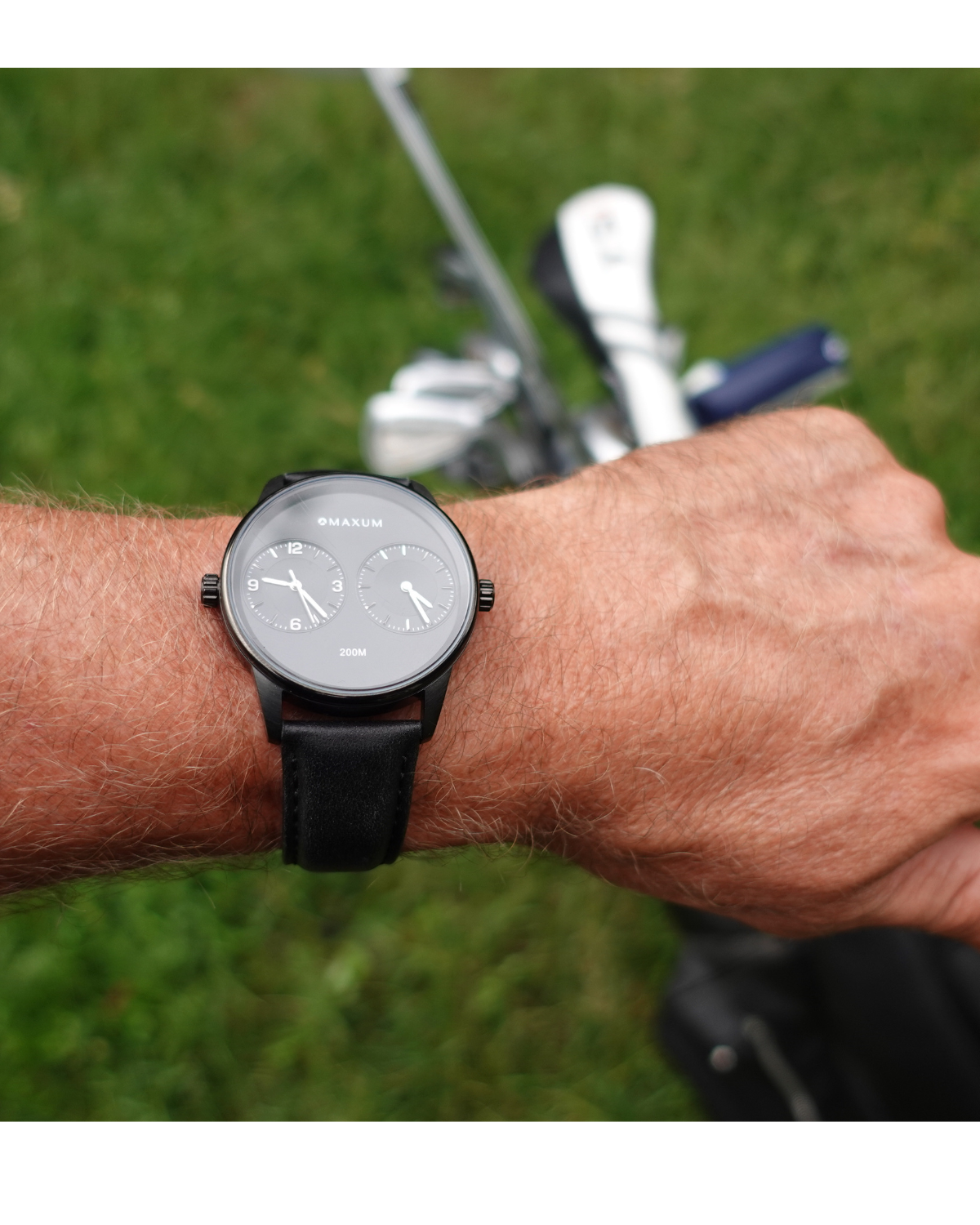 Maxum x Taylor Steele collab- black analog watch with a black dial, black case, and black leather band, branded with 'MAXUM' on the top and indicating '200M' water resistance. Person wearing  on a golf course