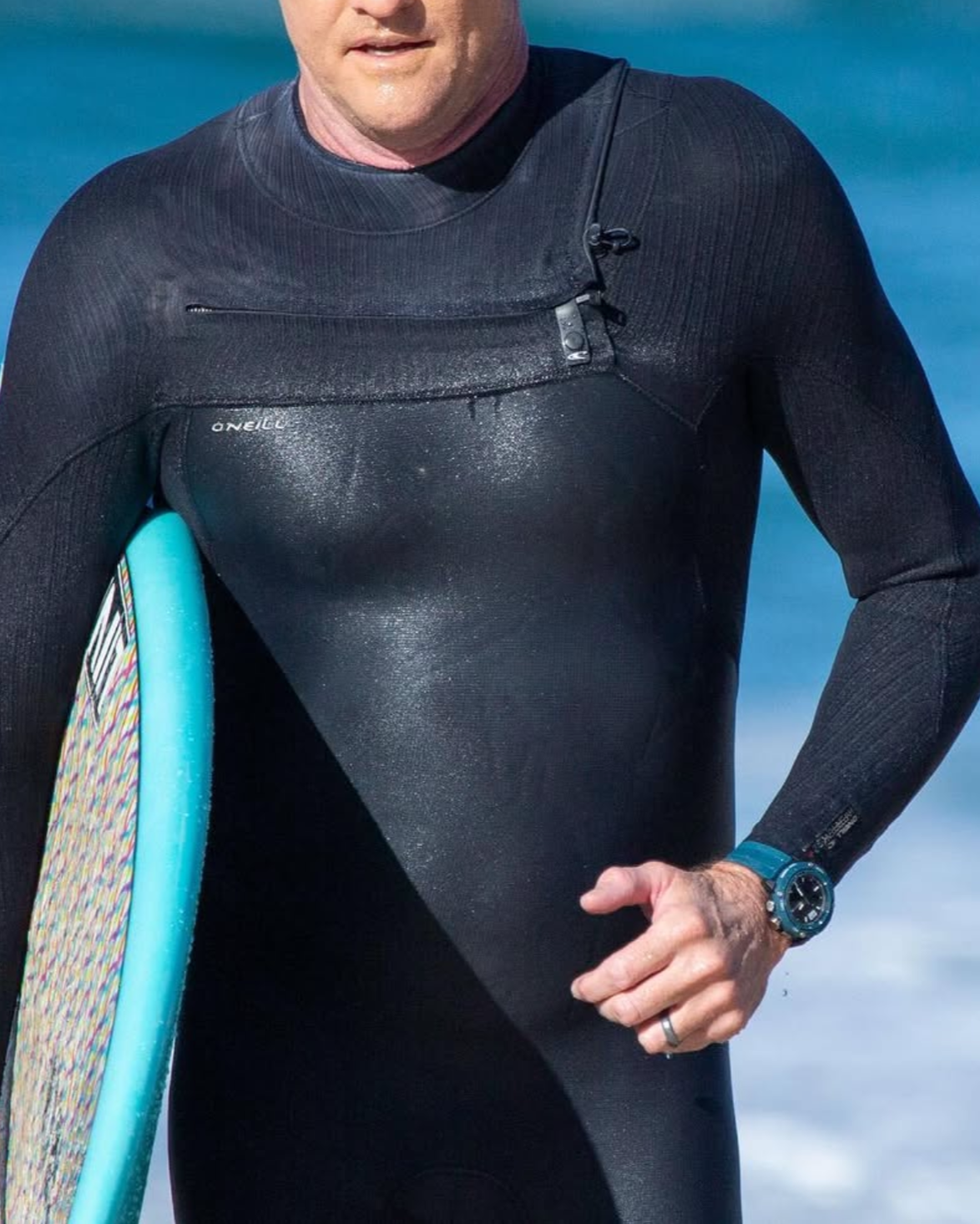 Maxum blue Conquest Analogue Digital Watch with a silicone strap and a digital display. On wrist of man coming out of surf in wetsuit with surfboard under arm