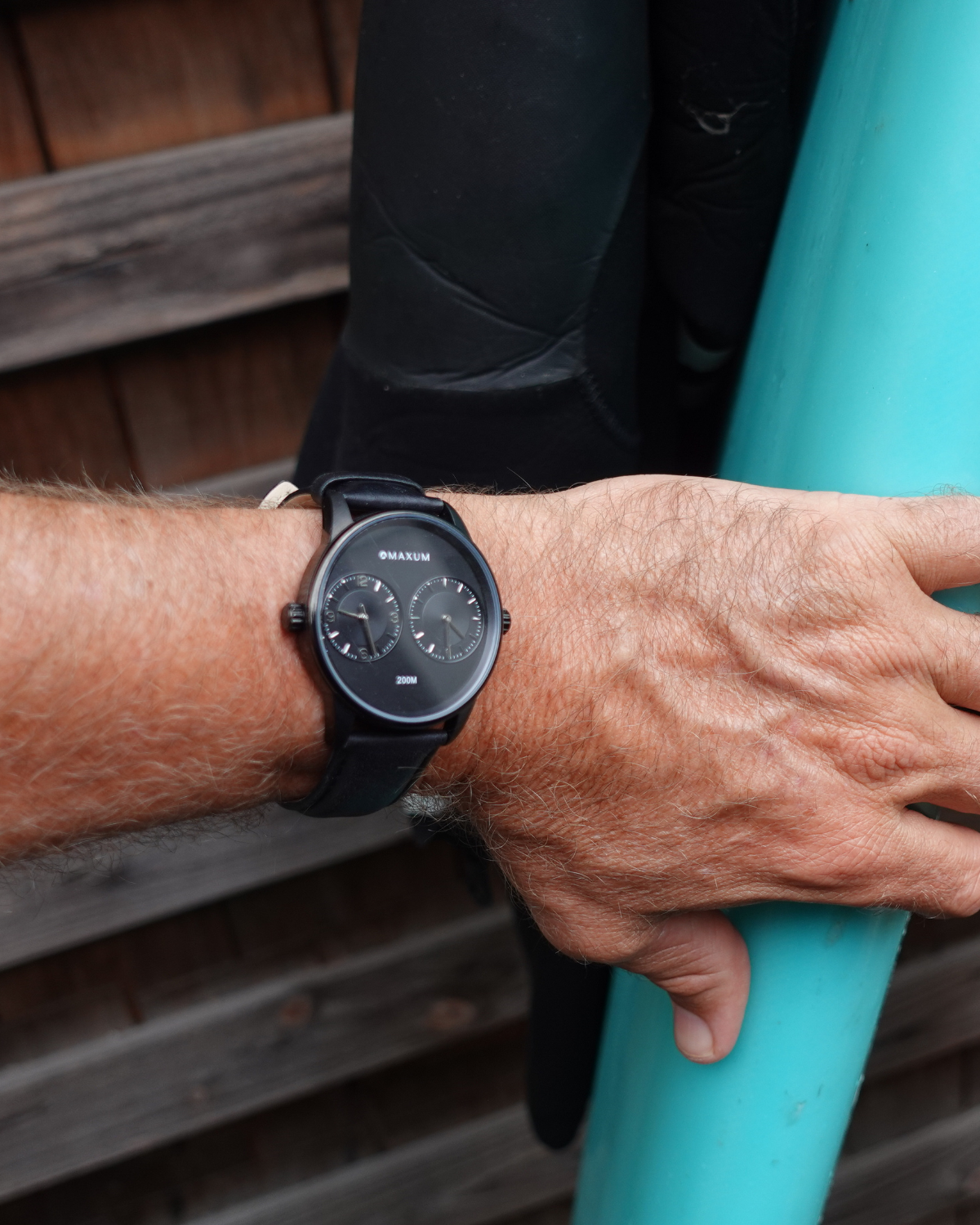 PMaxum x Taylor Steele collab- black analog watch with a black dial, black case, and black leather band, branded with 'MAXUM' on the top and indicating '200M' water resistance.. Worn by person holding a teal surfboard on a wooden background