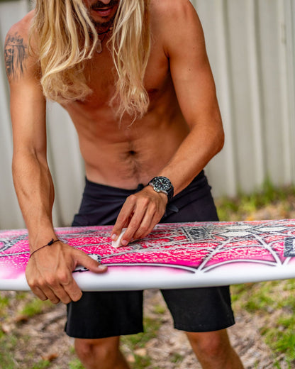 Kyuss King with long blonde hair holding a pink and white surfboard wearing Maxum Prince silver-tone casing watch with a black dial and black silicone strap, indicating 200m water resistance and featuring a date display.