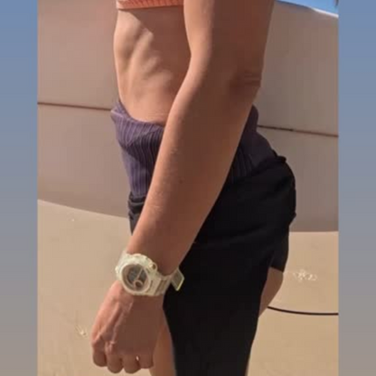 Maxum Endurance digital watch with a clear band and case, featuring a rose gold and white digital display, and buttons for various functions. Worn by female holding surfboard at beach 